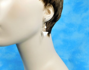 May include: A drop earring featuring a small white spherical bead suspended from a thin gold-toned metal wire, which connects to a flat, white triangular pendant. The earring hangs from a simple hook mechanism, showcasing a clean and geometric design.