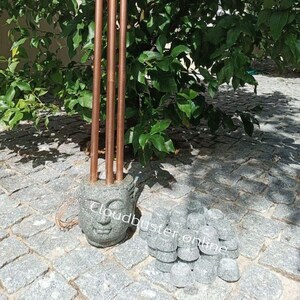 Könnte beinhalten: Ein Stein-Buddha-Kopf-Räucherstäbchenhalter mit vier kupfernen Räucherstäbchen. Der Halter ist von einem Haufen kleiner, runder, grauer Steine umgeben.