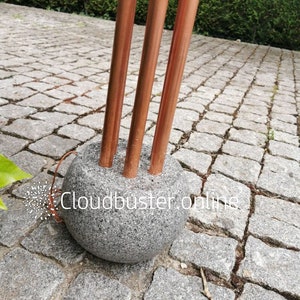 Op de afbeelding: Drie koperen buizen zijn in een ronde, grijze stenen basis gestoken. De basis staat op een grijs grind oppervlak. De tekst "Cloudbuster.online" is zichtbaar op de basis.
