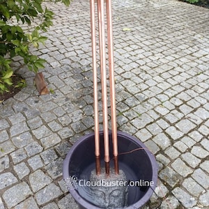 Könnte beinhalten: Ein Kupferbrunnen mit vier vertikalen Rohren, die sich von einer kreisförmigen Basis erstrecken. Die Rohre sind in einem dunkelgrauen Becken mit Wasser versenkt, wobei ein steinerner Buddha-Kopf sichtbar ist. Der Text "cloudbuster.online" befindet sich unten.