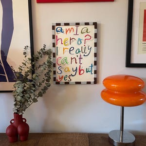 May include: A framed artwork with the text "am I a hero? I really can't say but yes" in colorful handwritten letters. A red vase with a unique shape holds greenery. An orange, double-layered lamp sits on a silver base.