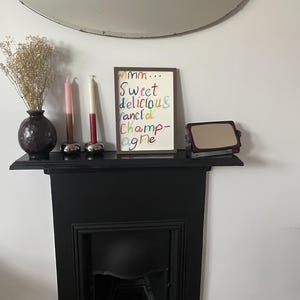 May include: A black fireplace mantel displays a framed artwork with colorful text that reads "Mmm... Sweet delicious ranci'd Champagne". A vase with dried flowers, two candles, and a small mirror are also on the mantel. A large, round mirror hangs above.