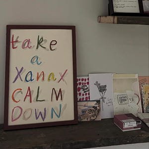 May include: A framed artwork with the words "take a Xanax CALM DOWN" in colorful, handwritten letters. The artwork is displayed on a wooden shelf alongside several greeting cards and a small box.