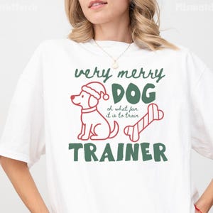 May include: A white t-shirt featuring a red outline of a dog wearing a Santa hat, a red striped bone, and green text that reads very merry DOG TRAINER oh what fun it is to train.