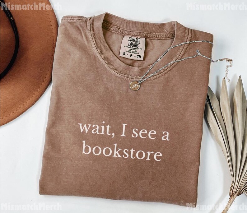 May include: A brown t-shirt with the text "wait, I see a bookstore" in white. A silver necklace with a pendant is on the shirt. A brown hat and dried leaves are in the background.