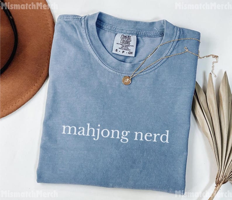 May include: A pale blue t-shirt with the words "mahjong nerd" printed in white. The shirt has a crew neck and short sleeves. A gold necklace with a pendant is draped across the shirt.