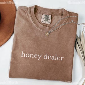 Op de afbeelding: Bruin t-shirt met de witte tekst "honey dealer". Het shirt heeft een ronde hals en een klein label op de kraag. Een zilveren ketting met hanger ligt over het shirt.