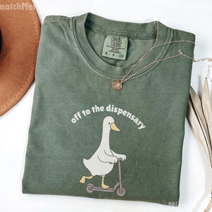 May include: An olive green t-shirt with the phrase "off to the dispensary" and a cartoon duck riding a scooter. A gold necklace with a pendant is draped across the shirt. The shirt is a Comfort Colors brand.