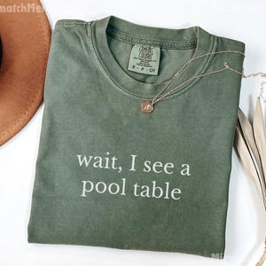 May include: A green t-shirt with the text "wait, I see a pool table" printed in white. The shirt is made of a soft, comfortable material.