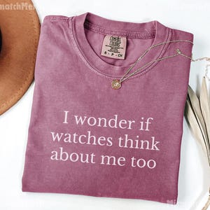 May include: A dusty rose Comfort Colors t-shirt with the text "I wonder if watches think about me too" in white. A gold necklace with a pendant rests on the shirt. A brown hat is visible in the upper left corner.