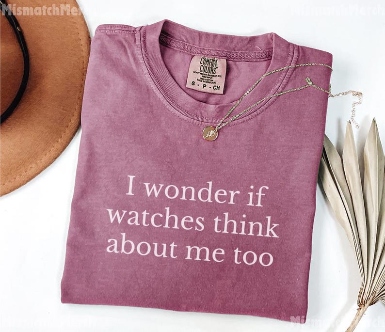 May include: A dusty rose Comfort Colors t-shirt with the text "I wonder if watches think about me too" in white. A gold necklace with a pendant rests on the shirt. A brown hat is visible in the upper left corner.