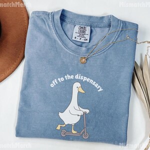 May include: A blue Comfort Colors t-shirt featuring a white duck riding a scooter, with the phrase "off to the dispensary" in white. A gold necklace and a brown hat are also visible.