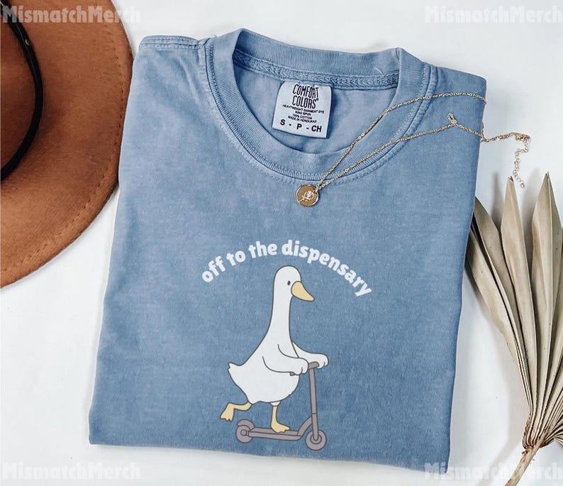 May include: A blue Comfort Colors t-shirt featuring a white duck riding a scooter, with the phrase "off to the dispensary" in white. A gold necklace and a brown hat are also visible.