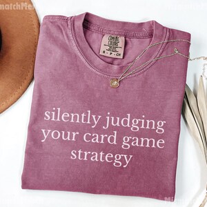 Peut inclure: Un t-shirt rose poussi&eacute;reux avec le texte blanc "silently judging your card game strategy". Un collier dor&eacute; avec un pendentif est pos&eacute; sur le t-shirt. Un chapeau marron et une plante s&eacute;ch&eacute;e sont en arri&egrave;re-plan.