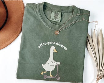 May include: An olive green t-shirt with the phrase "off to get a divorce" above a cartoon goose riding a scooter. The shirt has a crew neck and short sleeves. A gold necklace with a pendant is on the shirt.
