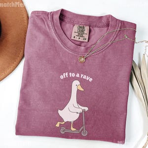 May include: A mauve Comfort Colors t-shirt with the phrase "off to a rave" and a cartoon goose riding a scooter. A gold necklace with a pendant rests on the shirt. The shirt is folded and displayed on a white surface.