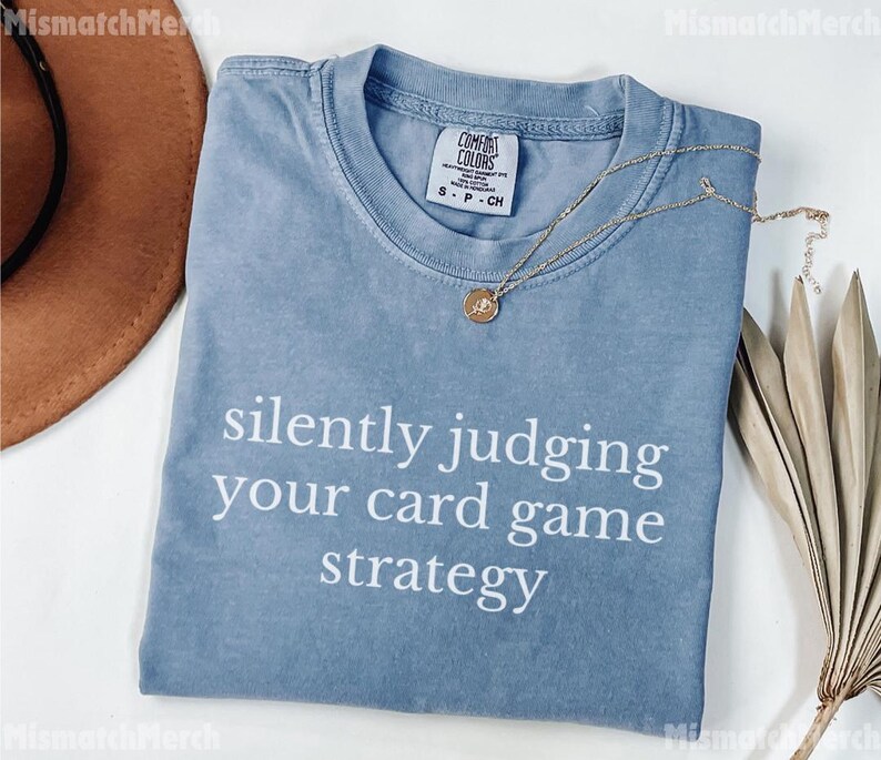 Peut inclure: Un t-shirt bleu Comfort Colors avec le texte "silently judging your card game strategy" en blanc. Un collier en or avec un pendentif est drap&eacute; sur le t-shirt. Un chapeau marron et une plante s&eacute;ch&eacute;e sont en arri&egrave;re-plan.