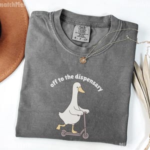 May include: A gray Comfort Colors t-shirt with the phrase "off to the dispensary" above a cartoon goose riding a scooter. The shirt has a crew neck and a small tag. A gold necklace is draped across the shirt.