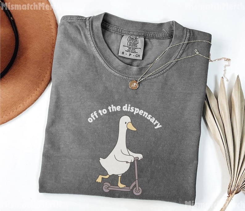 May include: A gray Comfort Colors t-shirt with the phrase "off to the dispensary" above a cartoon goose riding a scooter. The shirt has a crew neck and a small tag. A gold necklace is draped across the shirt.