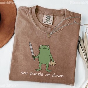 May include: A brown short-sleeved t-shirt featuring a green frog illustration holding a silver sword and a tan puzzle piece. The text "we puzzle at dawn" is printed below the frog. The shirt has a Comfort Colors brand label in the neck.