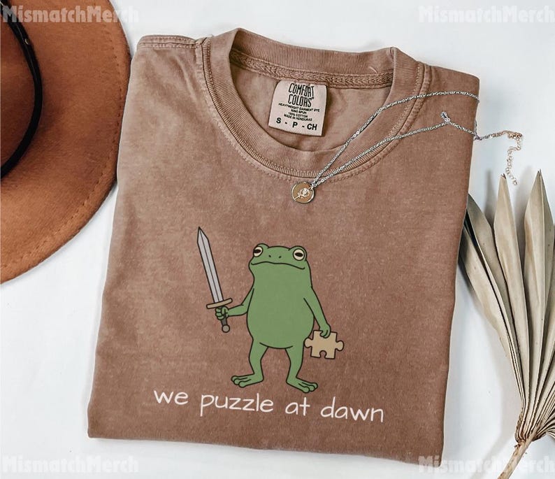 May include: A brown short-sleeved t-shirt featuring a green frog illustration holding a silver sword and a tan puzzle piece. The text "we puzzle at dawn" is printed below the frog. The shirt has a Comfort Colors brand label in the neck.