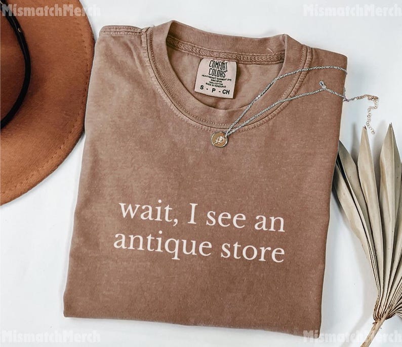 May include: A brown t-shirt with the text "wait, I see an antique store" in white. A silver necklace with a pendant is on the shirt. A brown hat is in the upper left corner.