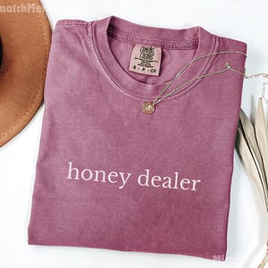 Op de afbeelding: Een stoffig roze t-shirt met de woorden "honey dealer" in het wit gedrukt. Een gouden ketting met een hanger ligt op het shirt. Het shirt heeft een ronde hals en een klein label.