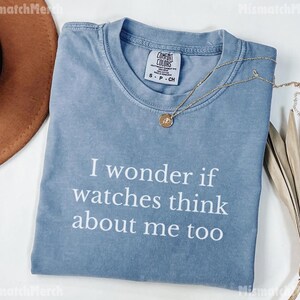 May include: A light blue Comfort Colors t-shirt with the text "I wonder if watches think about me too" in white. A gold necklace with a pendant is draped across the shirt. A brown hat is visible in the upper left corner.