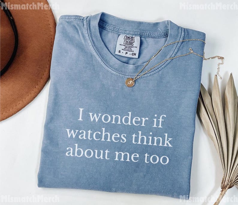 May include: A light blue Comfort Colors t-shirt with the text "I wonder if watches think about me too" in white. A gold necklace with a pendant is draped across the shirt. A brown hat is visible in the upper left corner.
