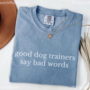 May include: A blue t-shirt with the text "good dog trainers say bad words" in white. The shirt has a crew neck and a small tag on the collar. A gold necklace with a pendant is draped across the shirt. A brown hat is visible in the background.