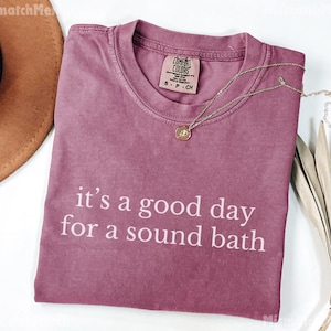 May include: A mauve Comfort Colors t-shirt with the phrase "it's a good day for a sound bath" in white lettering. A gold necklace with a pendant rests on the shirt. A brown hat and dried foliage are in the background.