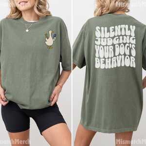 May include: An olive green short-sleeve t-shirt featuring a small graphic of a white duck holding a brown bone and a blue item on the front left chest. The back of the shirt displays the white text, "SILENTLY JUDGING YOUR DOG'S BEHAVIOR."