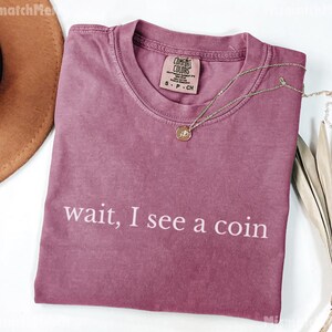 May include: A mauve Comfort Colors t-shirt with the phrase "wait, I see a coin" printed in white. A gold necklace with a pendant rests on the shirt. A brown hat is visible in the upper left corner.