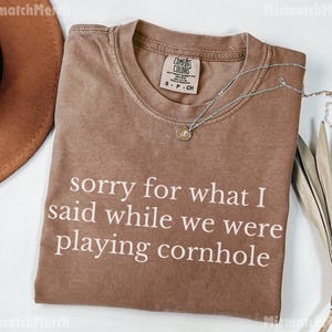 May include: A brown t-shirt with the text "sorry for what I said while we were playing cornhole" in white. A silver necklace with a pendant is on the shirt. The shirt is folded on a white surface.