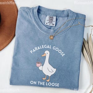 May include: A blue t-shirt featuring the text "PARALEGAL GOOSE ON THE LOOSE" and a cartoon goose with a coffee cup. The shirt has a crew neck and short sleeves. A gold necklace is draped across the shirt.