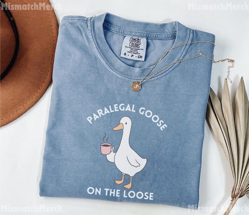 May include: A blue t-shirt featuring the text "PARALEGAL GOOSE ON THE LOOSE" and a cartoon goose with a coffee cup. The shirt has a crew neck and short sleeves. A gold necklace is draped across the shirt.