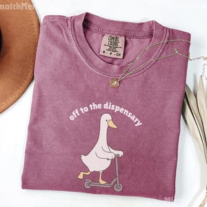 May include: A dusty rose t-shirt with the phrase "off to the dispensary" above a cartoon goose riding a scooter. The shirt has a crew neck and a small tag. A gold necklace with a pendant is draped across the shirt.