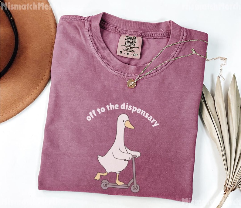 May include: A dusty rose t-shirt with the phrase "off to the dispensary" above a cartoon goose riding a scooter. The shirt has a crew neck and a small tag. A gold necklace with a pendant is draped across the shirt.