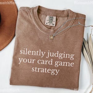 Peut inclure: Un t-shirt marron avec le texte "silently judging your card game strategy" en blanc. Le t-shirt a un col rond et des manches courtes. Un collier argent&eacute; avec un pendentif est sur le t-shirt. Un chapeau marron et une plante s&eacute;ch&eacute;e sont en arri&egrave;re-plan.