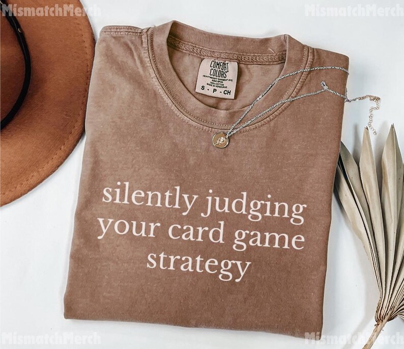 Peut inclure: Un t-shirt marron avec le texte "silently judging your card game strategy" en blanc. Le t-shirt a un col rond et des manches courtes. Un collier argent&eacute; avec un pendentif est sur le t-shirt. Un chapeau marron et une plante s&eacute;ch&eacute;e sont en arri&egrave;re-plan.