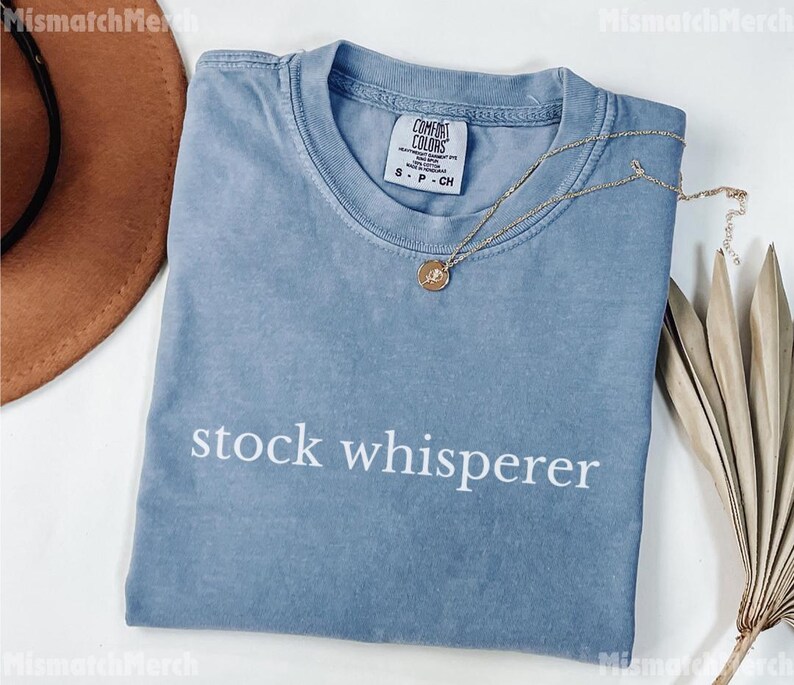 Puede incluir: Una camiseta azul claro con las palabras "stock whisperer" impresas en blanco. Un collar dorado con un colgante est&aacute; drapeado sobre la camiseta. La camiseta est&aacute; doblada sobre una superficie blanca.