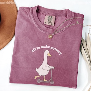 May include: A dusty rose Comfort Colors t-shirt with the phrase "off to make pottery" and a cartoon goose riding a scooter. A gold necklace with a pendant is draped on the shirt. A brown hat and dried plant are in the background.
