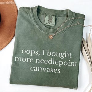 May include: An olive green t-shirt with the text "oops, I bought more needlepoint canvases" in white. A gold necklace with a pendant rests on the shirt. A brown hat and dried plant are in the background.