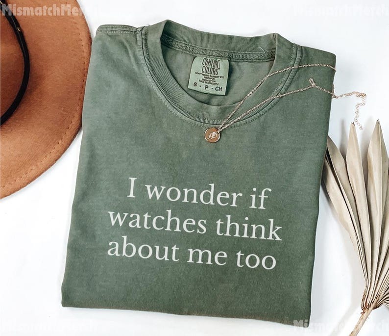 May include: An olive green t-shirt with the text "I wonder if watches think about me too" in white. A gold necklace with a pendant is draped across the shirt. A brown hat and dried leaves are in the background.