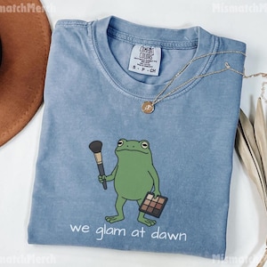 May include: A blue short-sleeved t-shirt featuring a graphic of a green frog holding a makeup brush and a palette. The text "we glam at dawn" is printed below the frog. The shirt has a Comfort Colors label at the neck and a crew neckline.