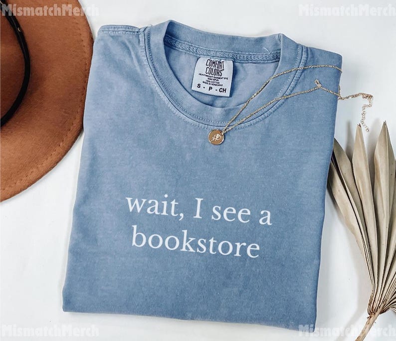 May include: A blue t-shirt with the text "wait, I see a bookstore" in white. The shirt has a crew neck and short sleeves. A gold necklace with a pendant is draped across the shirt.