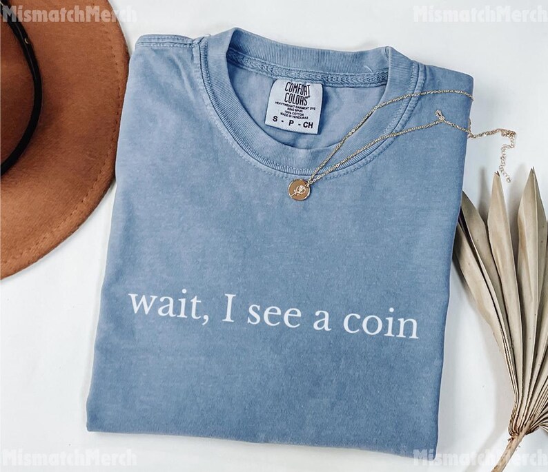 May include: A blue t-shirt with the phrase "wait, I see a coin" printed in white. The shirt has a crew neck and short sleeves. A gold necklace with a pendant is draped on the shirt.