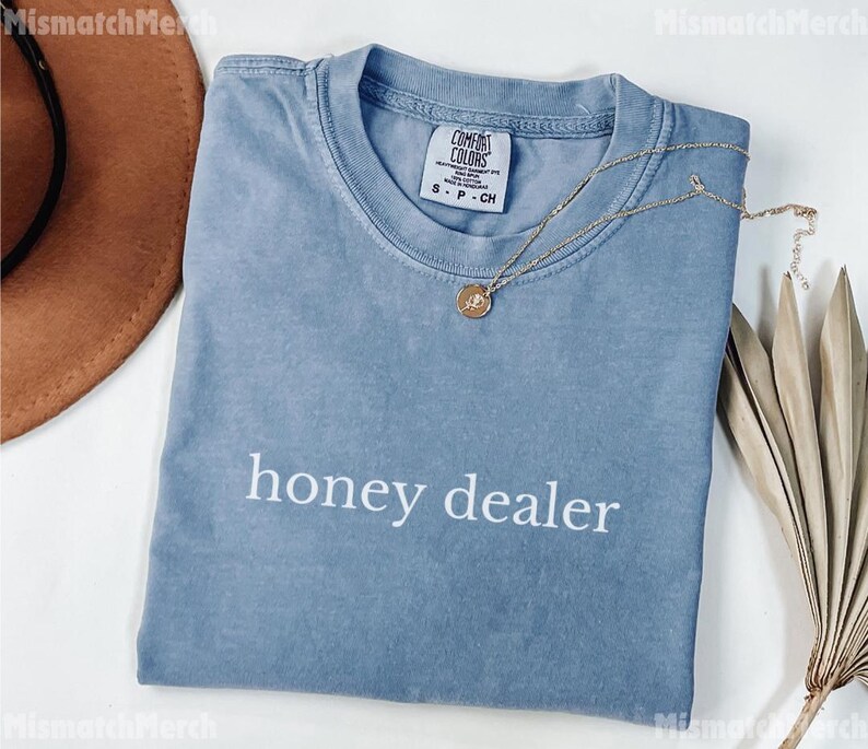 Op de afbeelding: Een blauw t-shirt met de woorden "honey dealer" in het wit gedrukt. Een gouden ketting met een hanger ligt op het shirt. Het shirt is opgevouwen en heeft een Comfort Colors-label.