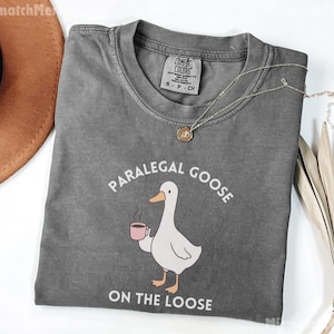 May include: A grey t-shirt featuring the text "PARALEGAL GOOSE ON THE LOOSE" and a cartoon goose with a coffee cup. The shirt has a crew neck and short sleeves. A gold necklace is draped across the shirt.
