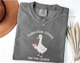 May include: A gray t-shirt with the text "PARALEGAL GOOSE ON THE LOOSE" and a cartoon goose holding a coffee cup. The shirt has a crew neck and short sleeves. A gold necklace is draped across the shirt.
